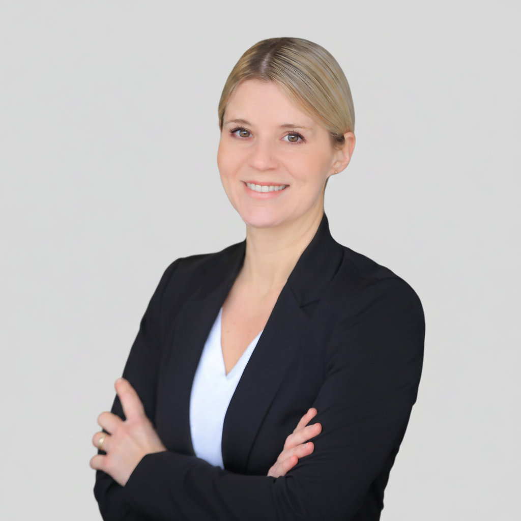The image shows a woman with blonde hair, wearing a black blazer over a white top. She is smiling and has her arms crossed. The background is plain and light-colored.