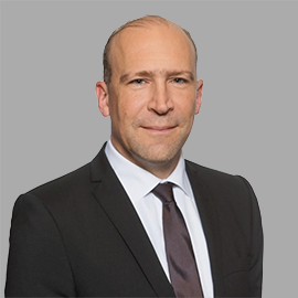 The image shows a person wearing a formal suit with a black jacket, white shirt, and dark tie.