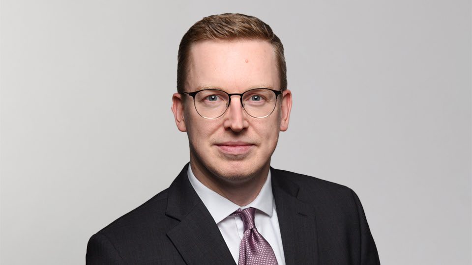 The image shows a person wearing a dark suit with a white shirt and a patterned tie.