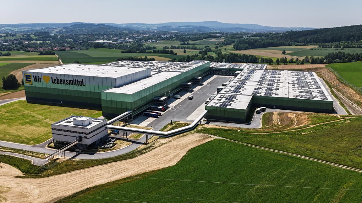 The image depicts a large industrial complex, specifically the logistics hub for the company Lidl. The facility consists of multiple expansive warehouse buildings with a modern design, featuring a flat roof and a green color that blends with the surrounding agricultural landscape. The grounds around the warehouse have been cultivated and are bordered by vast fields, illustrating the rural setting of the logistics center.

**Key Features Noticed in the Image:**
1. **Branding**: Visible branding of "Lidl" on the facility indicates the ownership or the purpose of this building.
2. **Infrastructure**: Includes multiple roads within the complex for vehicular access, such as trucks likely used for logistics and distribution.
3. **Surroundings**: The complex is situated in a picturesque, countryside setting with lush greenery and rolling hills visible in the background, reflecting its integration into a less urbanized area.
4. **Structure Layout**: The layout is spread out with a spacious design for logistical operations, including loading docks and expansive roof areas.
5. **Adjacent Buildings**: There appears to be a smaller auxiliary building connected by a bridge or walkway, serving as an office or support structure for the main warehousing operations.

This image overall portrays a modern, efficient logistical hub specifically tuned for the operations of Lidl, focusing on warehousing, distribution processes, and set in a landscape that balances industrial utility with natural aesthetics.