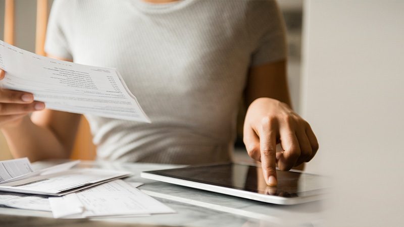 The image shows a person engaged in a task involving paperwork and a tablet. 

**Elements in the Image:**

1. **Person in Gray**: 
   - **Presence**: A person wearing a gray t-shirt is visible, focusing on the task at hand.
   - **Activity**: They are holding papers while interacting with a tablet.

2. **Paperwork**: 
   - **Description**: A hand is holding sheets of paper, likely bills or documents.
   - **Function**: Appears to be engaged in reviewing, organizing, or processing.

3. **Tablet Device**:
   - **Description**: Positioned on a table, the person is using their finger to interact with the touchscreen.
   - **Possible Use**: Could be used for recording data, research, or payment transactions.

4. **Table Setting**:
   - **Surroundings**: Documents scattered around imply active engagement in managing or processing information.

**Contextual Interpretation**:

- **Scenario**: The image likely depicts someone balancing digital and physical records, perhaps for financial management, research, or administrative purposes.

This could also illustrate an environment where traditional documentation meets modern technology, indicating productivity or multitasking in the professional or personal space. 

Such a setting underscores the blending of digital and analog methods in everyday tasks, showcasing a modern work dynamic.