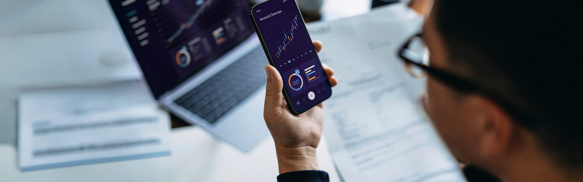 The image shows a person holding a smartphone displaying graphs and data, while a laptop on a desk also shows similar digital data visualizations and graphs. Several paper documents with printed information are scattered around the laptop.